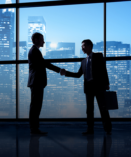 Photograph of two men in suits shaking hands with dark silhouettes, and in the background, windows with buildings in the night